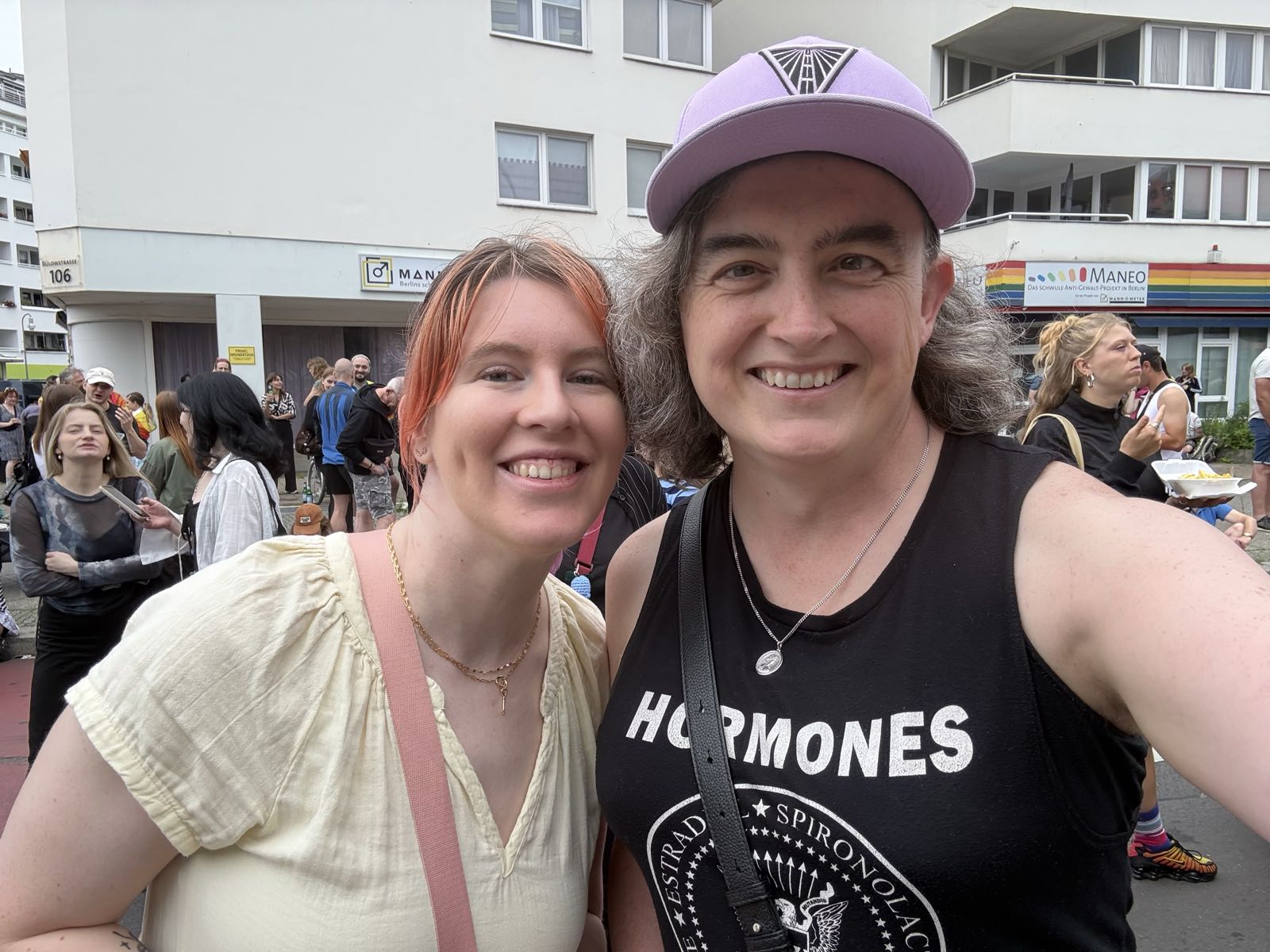 Two smiling women look at the camera. One has orange hair and is wearing a light yellow sundress, and one is in a black muscle-T with a lavender baseball cap on.