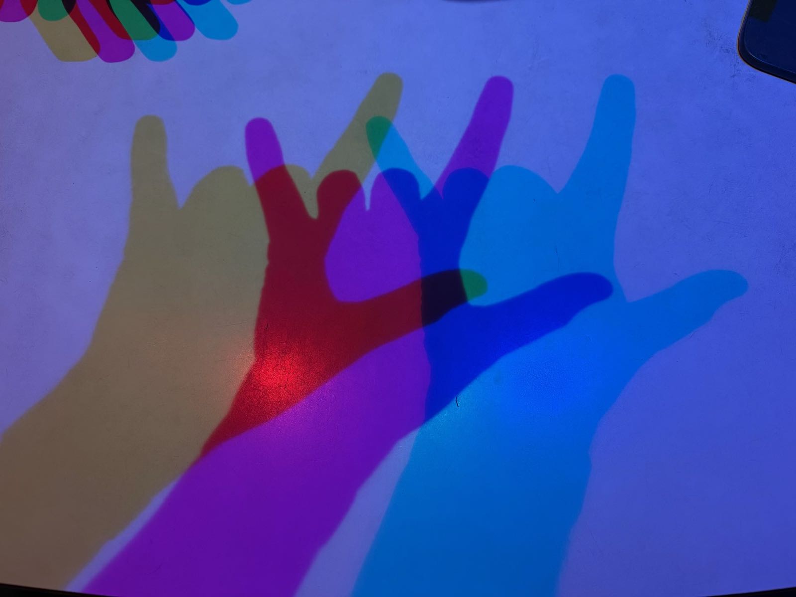 Several shadows of different colors are cast by a hand (off-camera) held in the ASL for “I love you” (🤟).