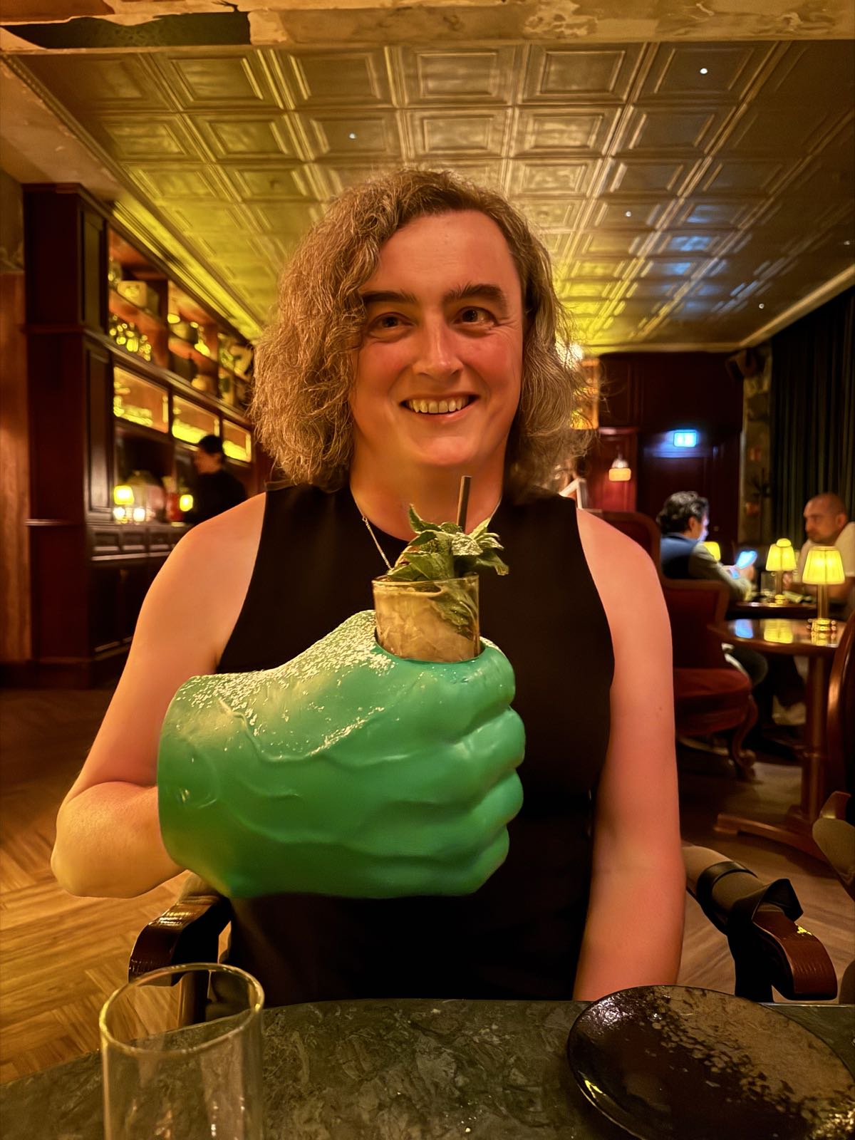 A woman in a black sleeveless dress sits at a table. She’s holding a cocktail. The cocktail is housed inside a large, green hand, so it appears that the woman’s right hand has swollen to many times its actual size.