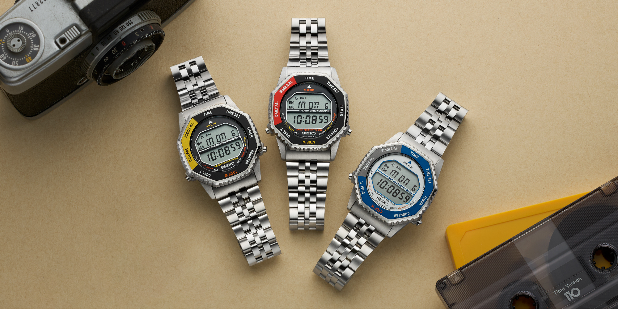 Three digital wristwatches sit on a brown table. They are identical except for their dial and bezel colors. One is black and yellow, one black and red, and the last is blue and gray.