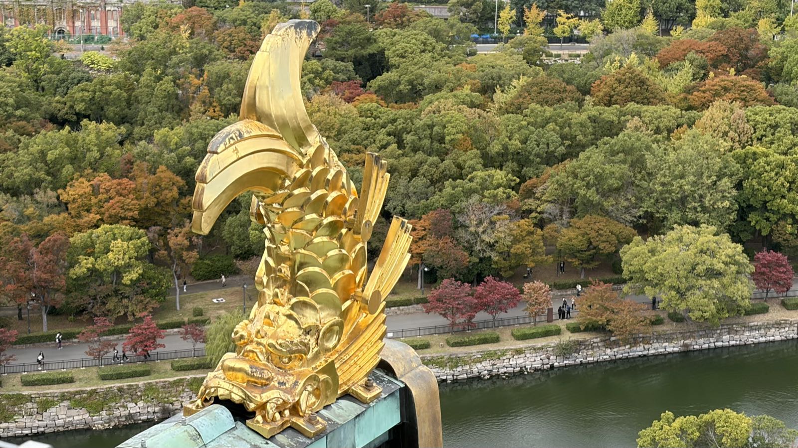 A stylized golden fish stands at the edge of a roof, high above autumn trees and a body of water.