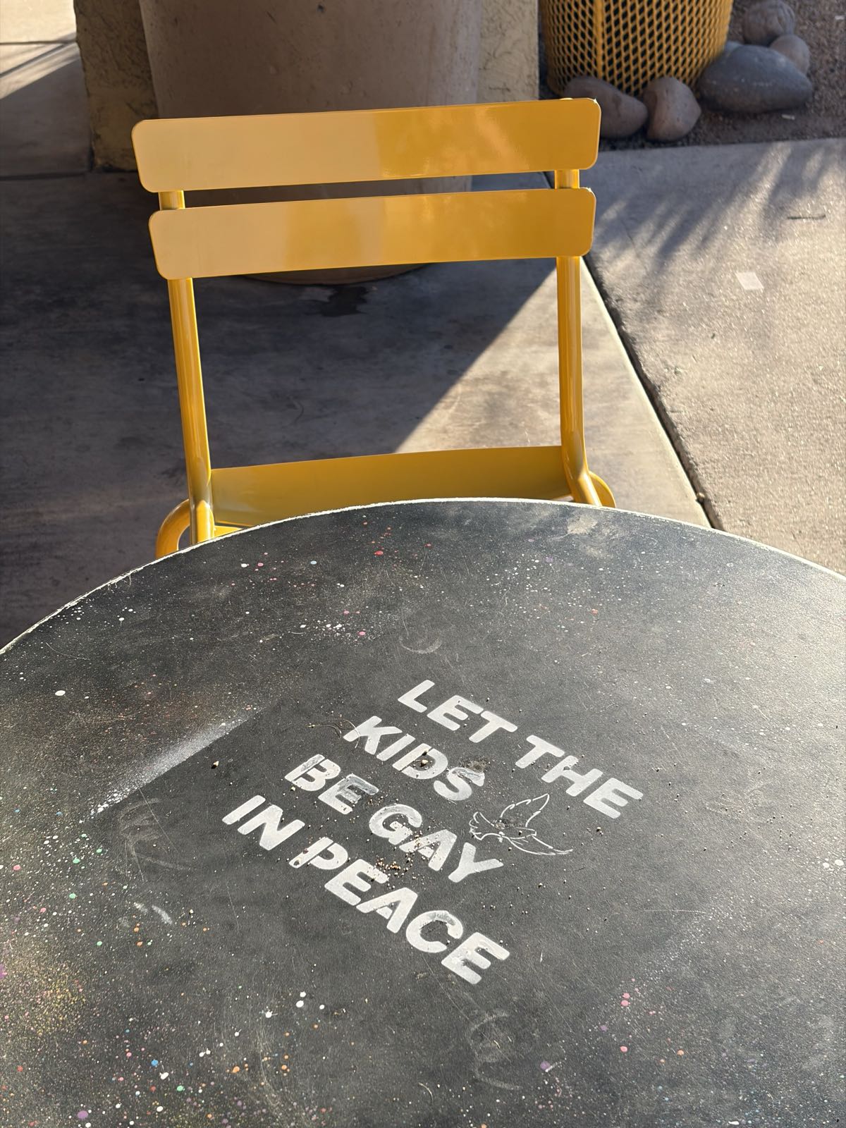 A dark, round table at which one yellow chair sits. On the table, the words “LET THE KIDS BE GAY IN PEACE” are stenciled.