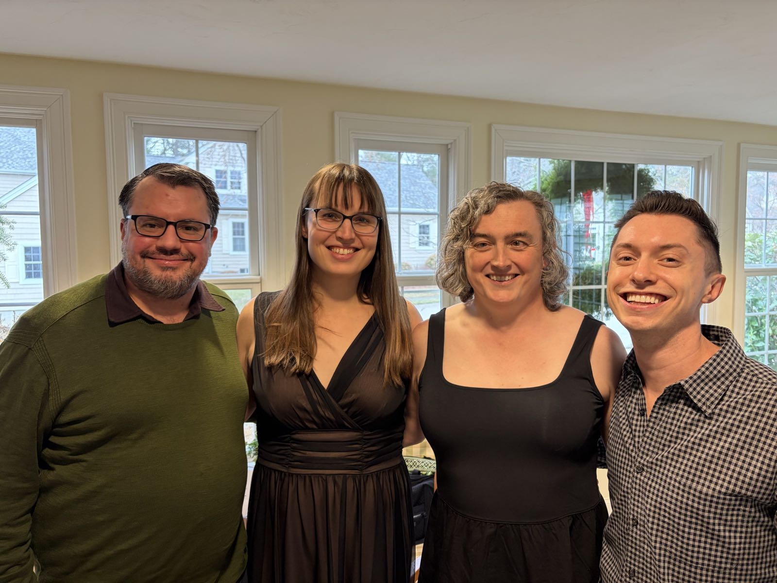 Two men and two women pose together for a photo. The man on the left is wearing a green sweater. To his right is a woman in a brown dress, then a woman in a black dress, and finally a man in a checkered button-up.