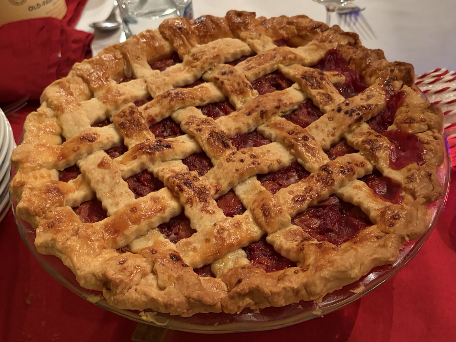 A pie is sitting on a table. The pie’s top crust is done in a diagonal grid pattern with red filling peeking through between the slats.
