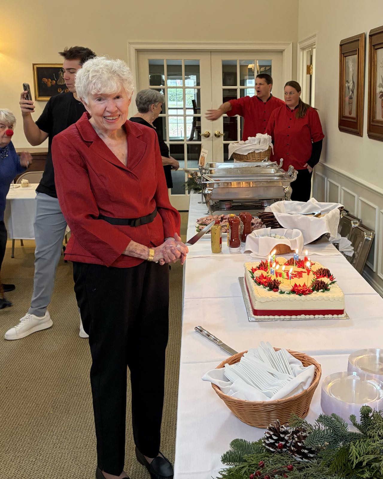 A woman in a red top and black pants smiles for the camera. In her hand is a large kitchen knife. There’s a sheet cake on the table in front of her.