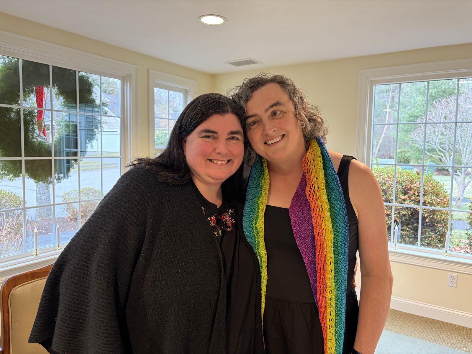 Two women smile for the camera. The one on the left is wearing black; the one on the right has a rainbow shawl over a black dress. They look alike.