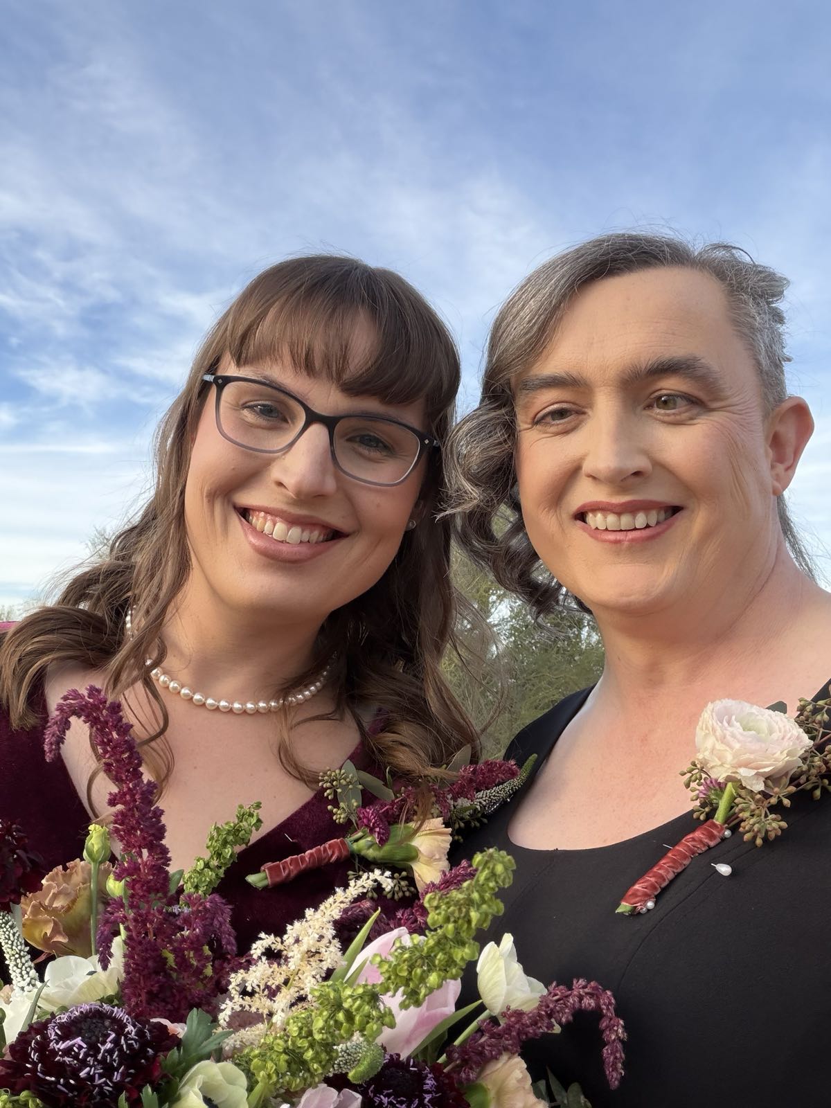 Two smiling women take a selfie together. They’re both wearing dresses, and each has a corsage pinned to her dress on the left. The woman on the left is holding a bouquet of flowers.