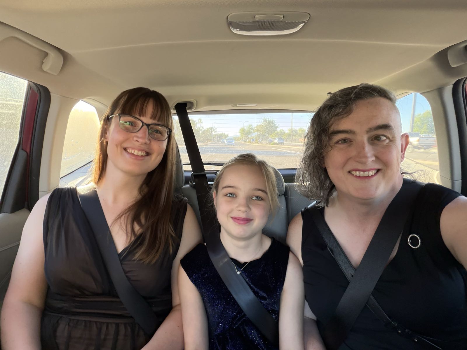 Two women and one girl are sitting in the backseat of a car. The woman on the left has long, straight, brown hair, and is wearing glasses. The woman on the right has wavy, asymmetric hair. The child in the middle has straight, dirty-blonde hair. All three of them are wearing dresses.