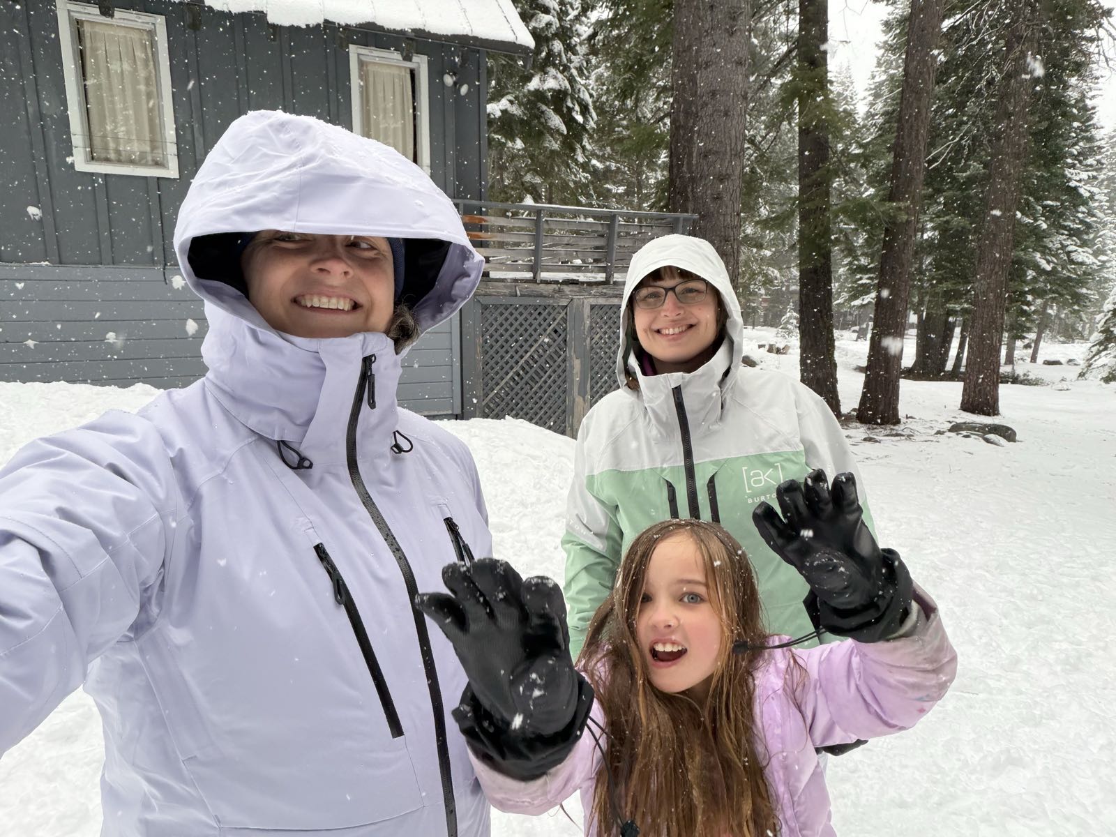 Two women and a girl, all in snow gear, pose for a selfie. The woman in a lavender snow jacket is taking the selfie. The other woman is in mint green, and the child is in pink, and has long hair. The child is making a very silly face at the camera.