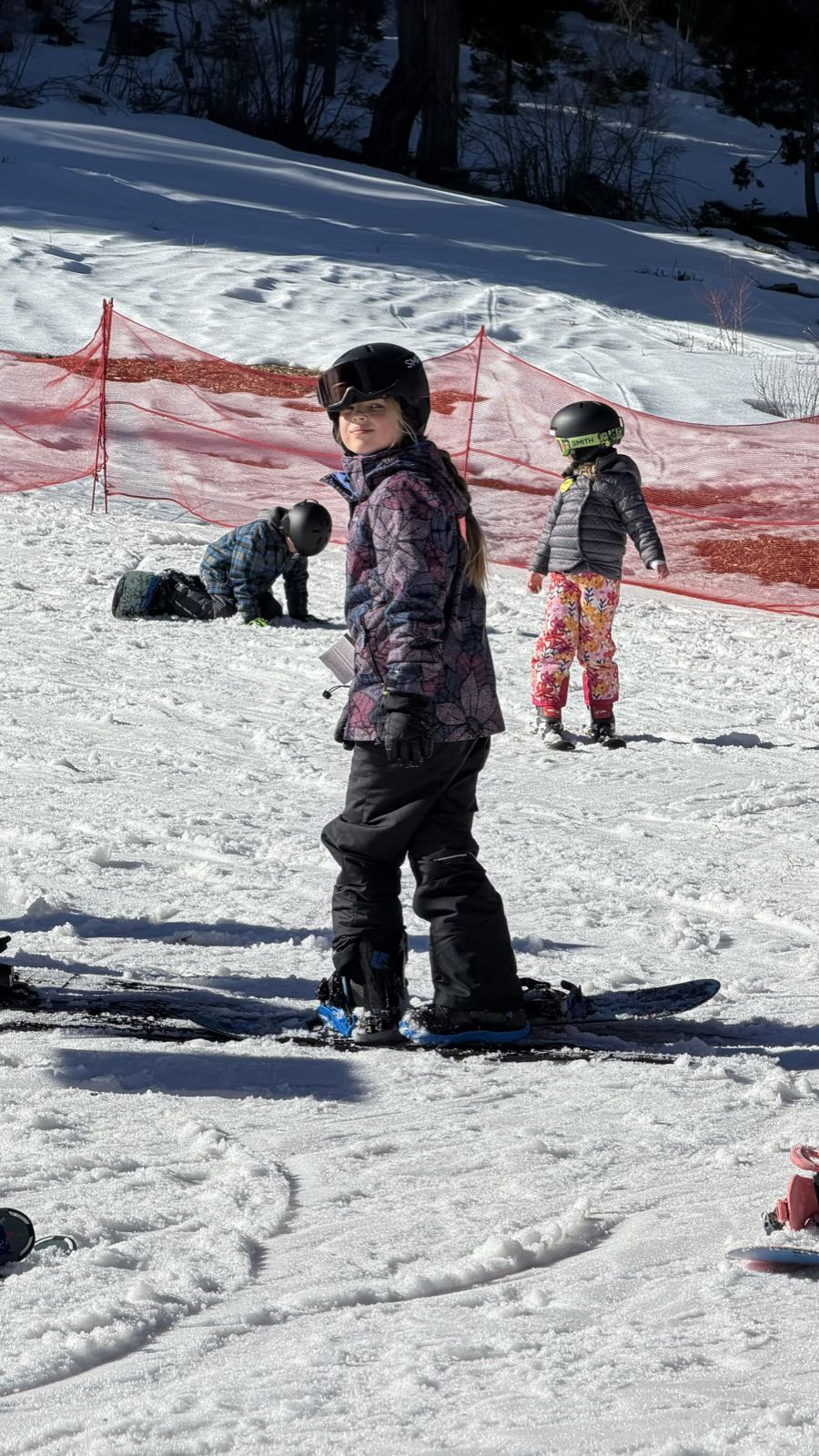 A child is standing on a snowboard. She’s wearing a purple ski jacket and black ski pants.