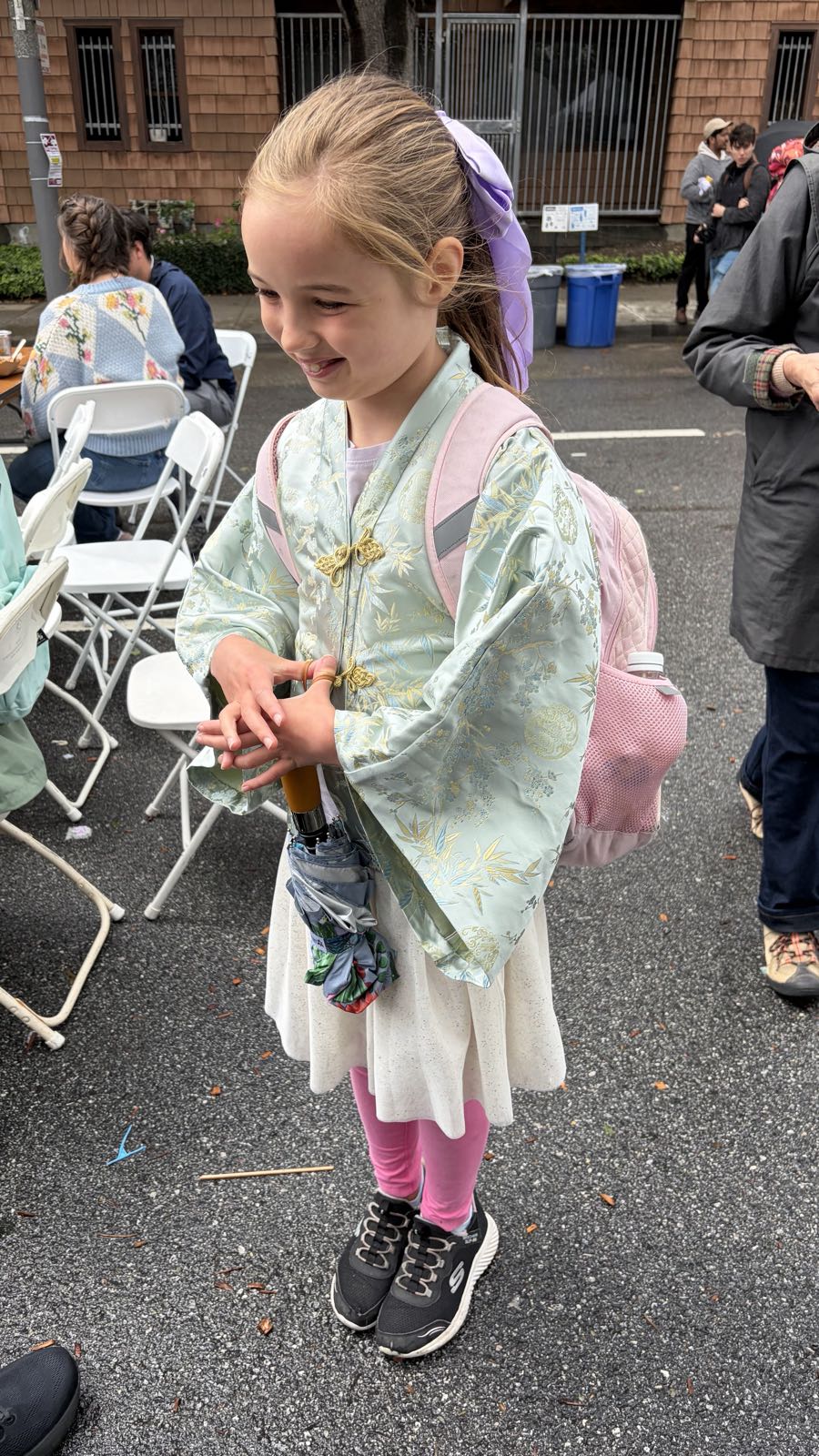 A child is smiling at somebody off-camera. She’s wearing a mint-green, Asian jacket over a white skirt, pink pants, and black shoes. She has a lavender-colored bow in her hair.