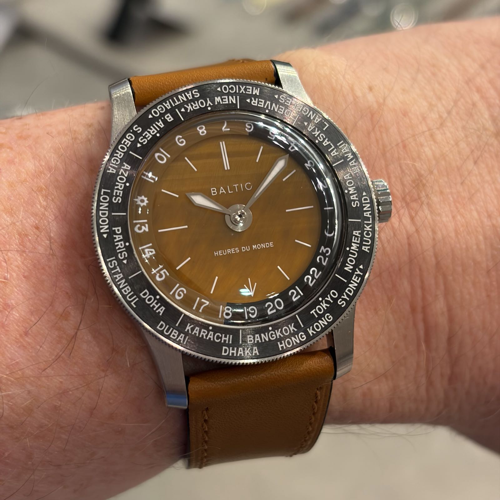 A watch with a brown dial and strap is on wrist. The watch’s black bezel lists many city names from around the world.