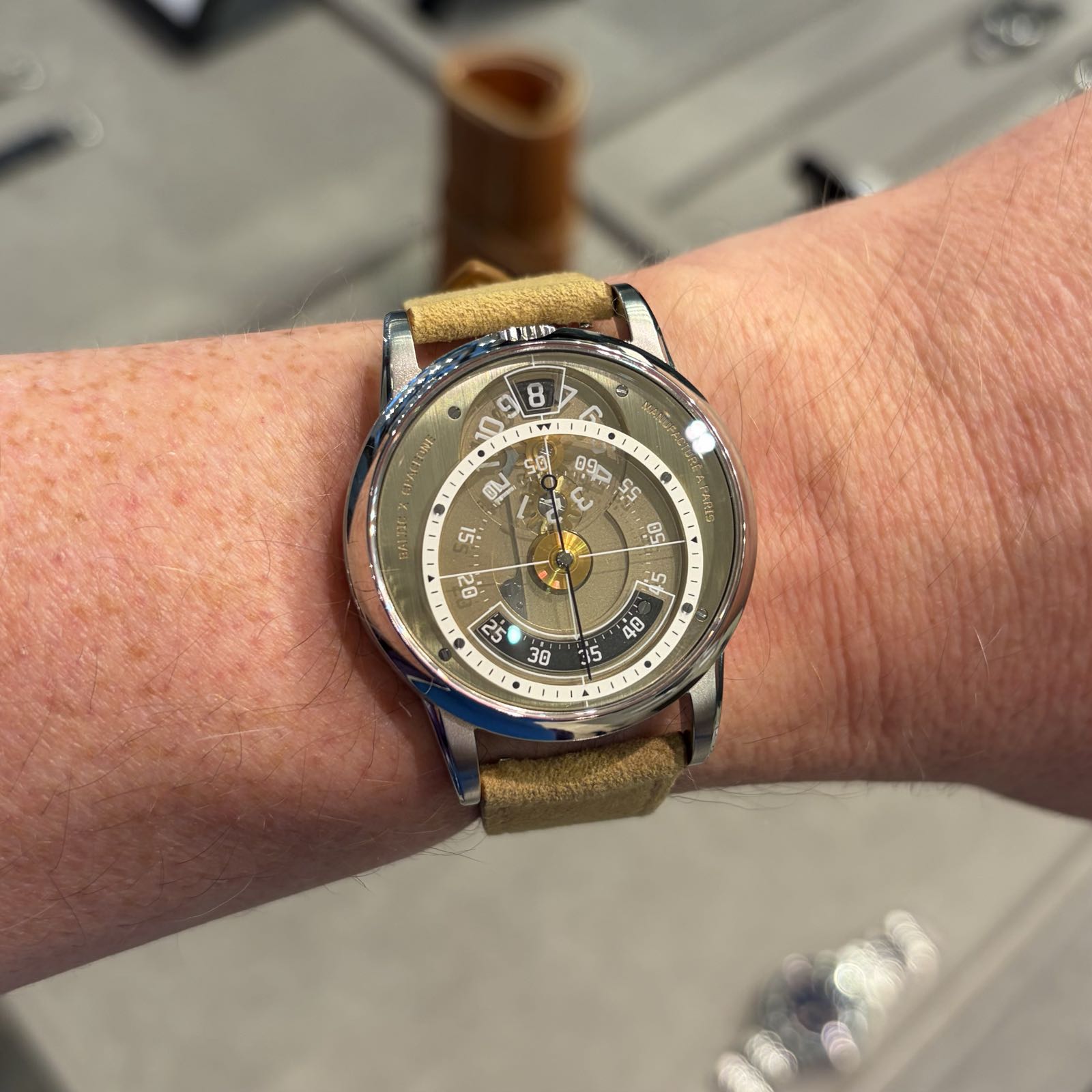 A very strange watch with a gold colored dial is being worn on a beige suede strap.