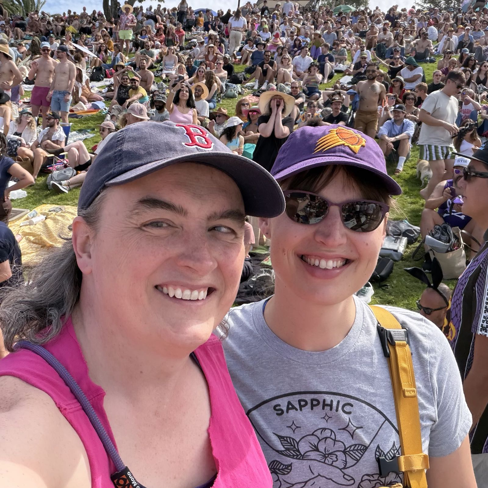 Two women smile at the camera for a selfie. They’re in a crowded park on a sunny day.