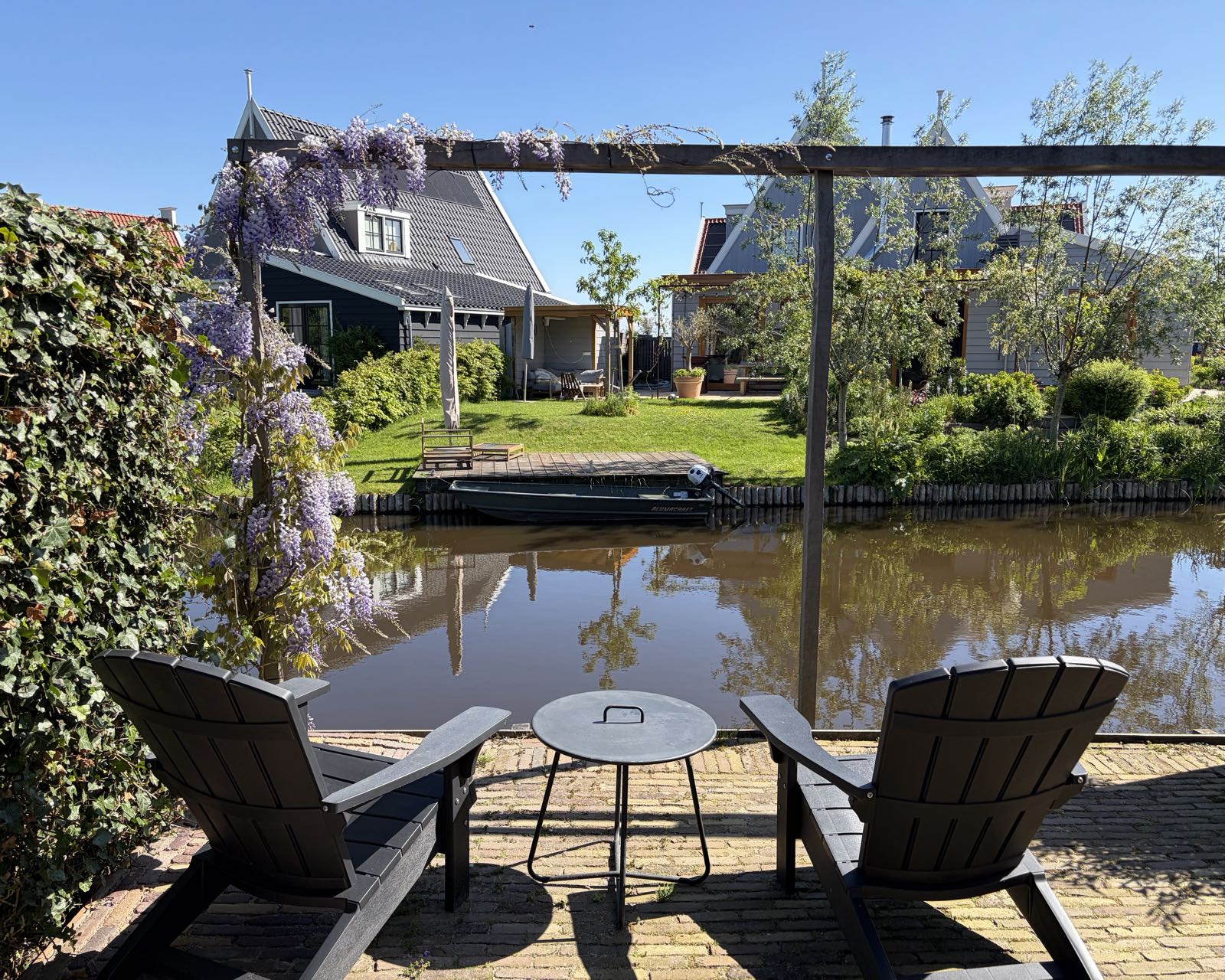 Two wooden chairs sit facing a small canal, with a table in between them. It’s a sunny day, and there are a couple of houses on the other side of the canal.
