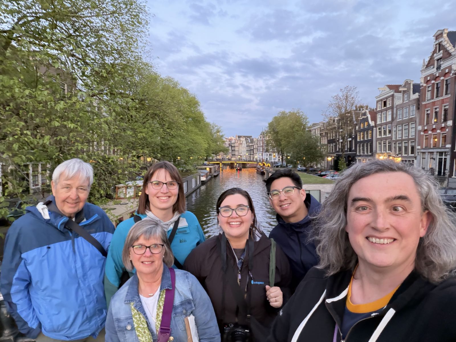 Six people pose for a photo. Behind them is a canal lined with townhouses. Everyone’s smiling for the camera. The person on the right has their arm extended, so they are presumably the photographer.