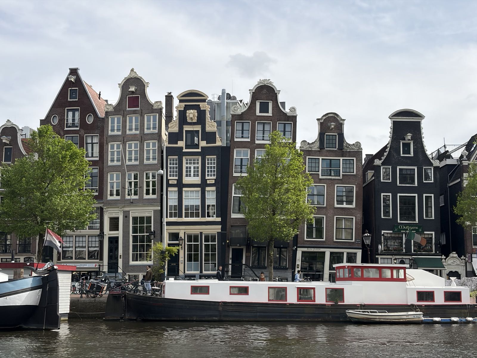 Six tall houses in Amsterdam are pressed up next to each other, all facing a canal. The houses are all somewhat askew, and appear to be holding each other up.