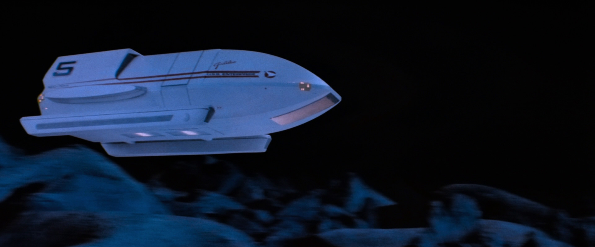 A spaceship is flying over rocky terrain. On the side of the spaceship, the number 5 is prominently printed.