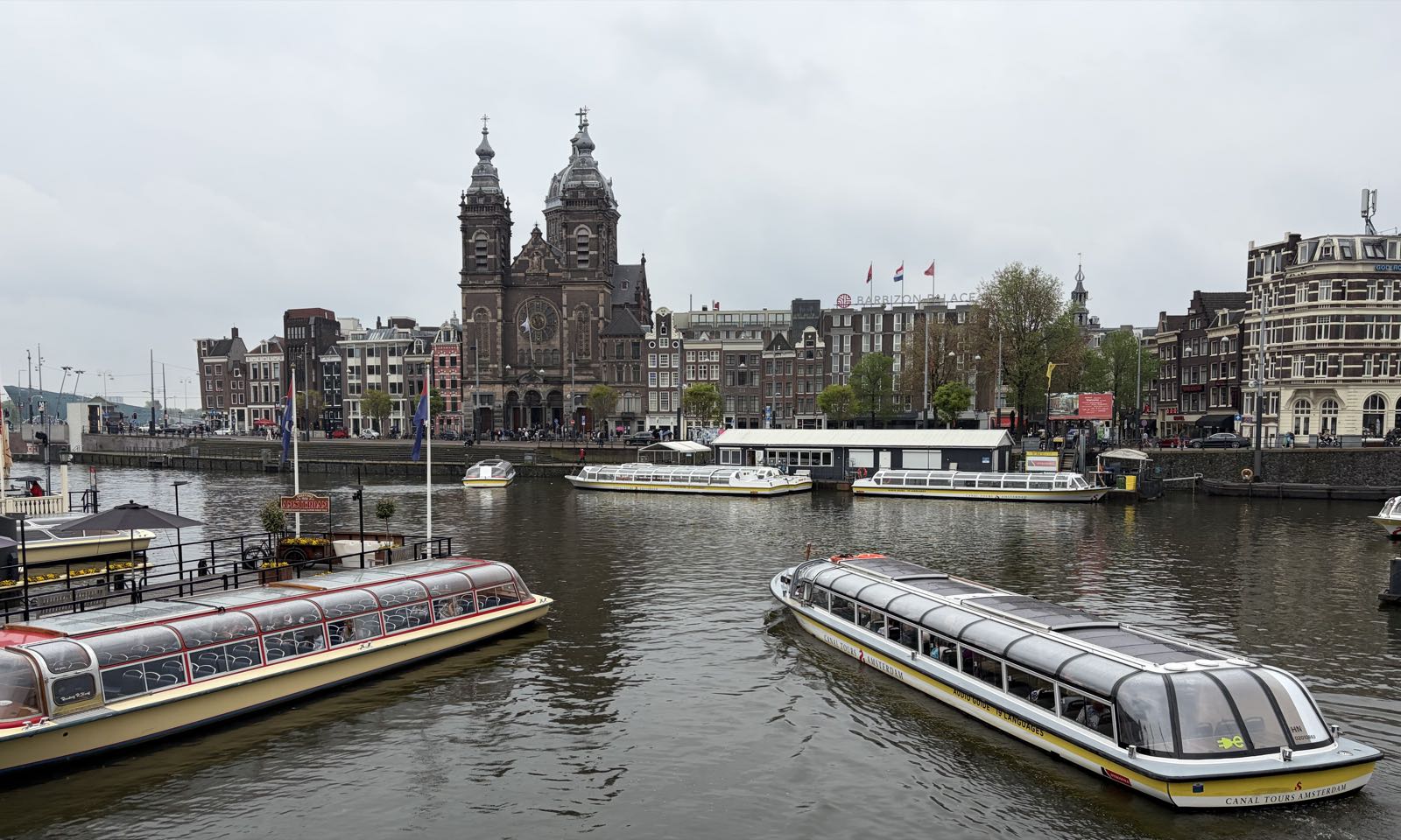 A brick cathedral stands on the far side of a river, surrounded by smaller city buildings. In the foreground are two passenger ferries.