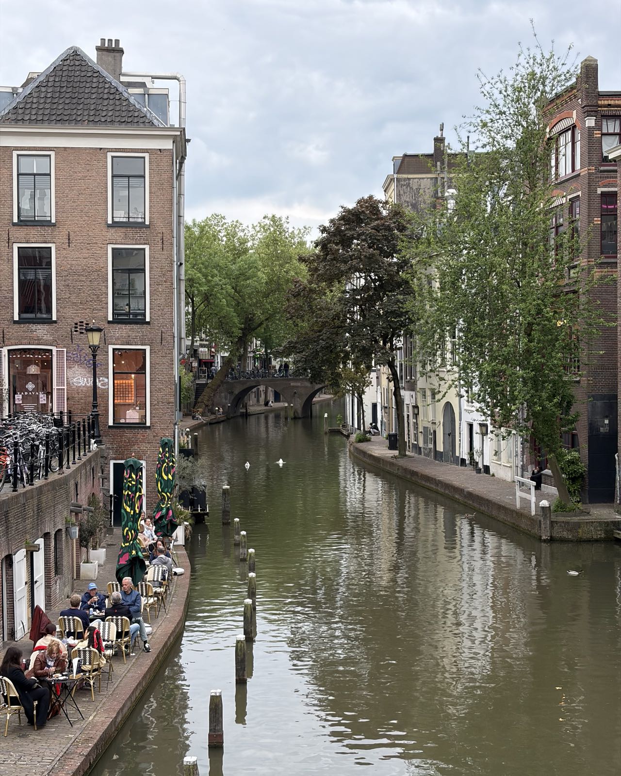 Many people sit at small cafe tables along the bank of a canal in a European-looking city.