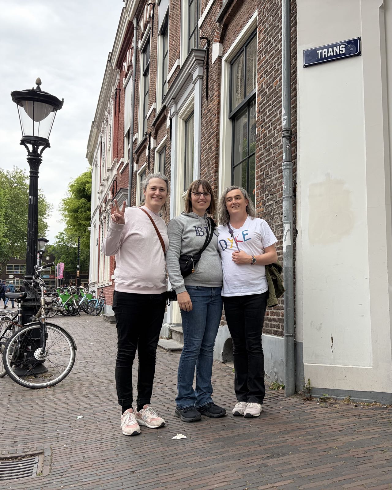 Three women pose for a photo in front of a row of buildings. Above them, the street sign reads “TRANS”.