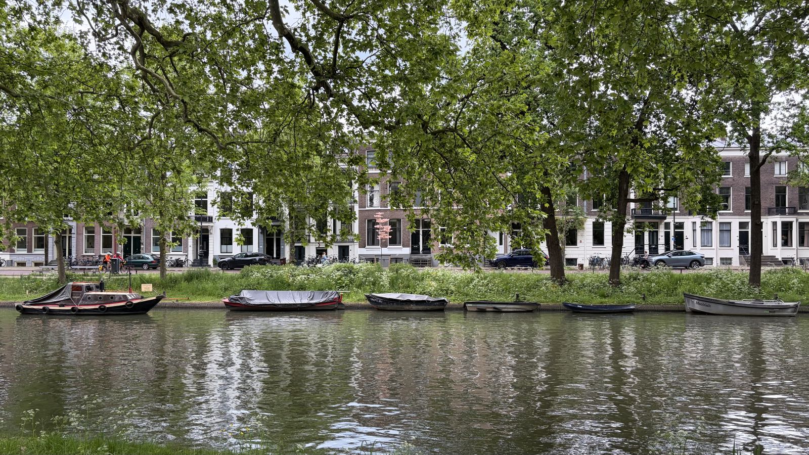 In the foreground is a canal. The far bank fo the canal has a number of boats tied up, then a row of trees, then a number of townhouses.