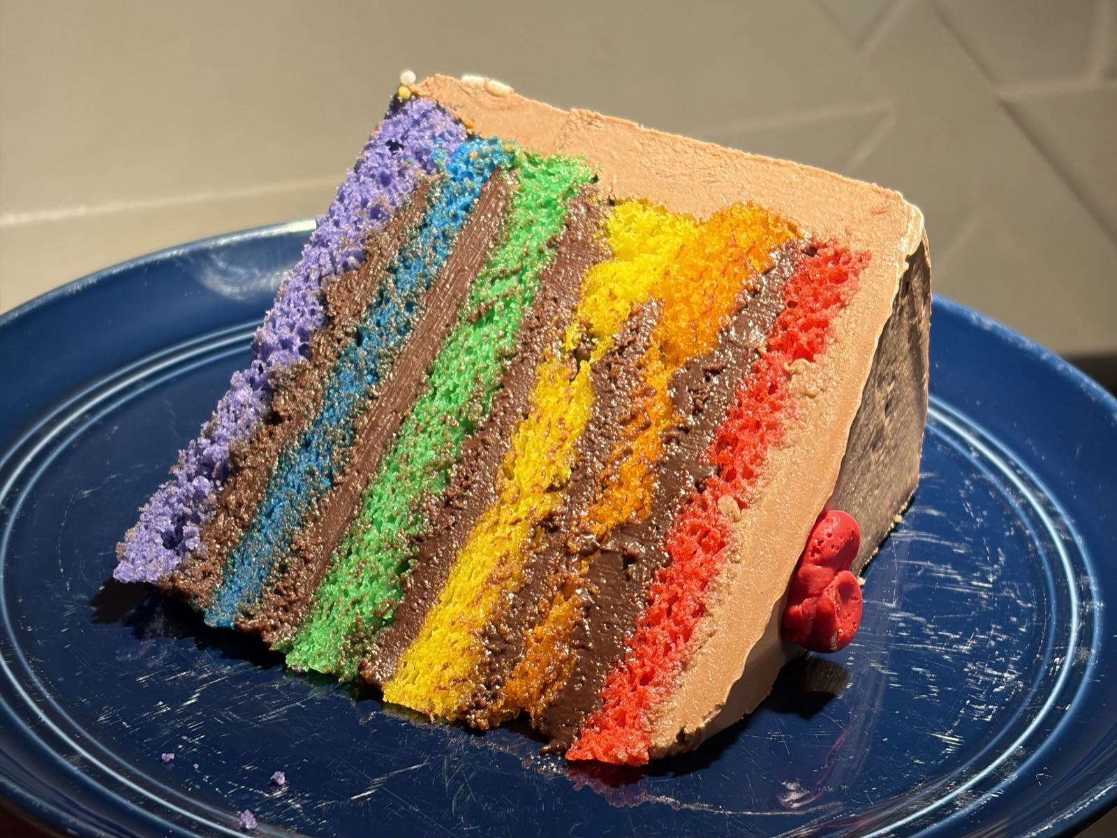 A slice of cake sits on a blue plate. The cake has six layers, each a color of the pride flag. The frosting between the layers and on the outside is chocolate.