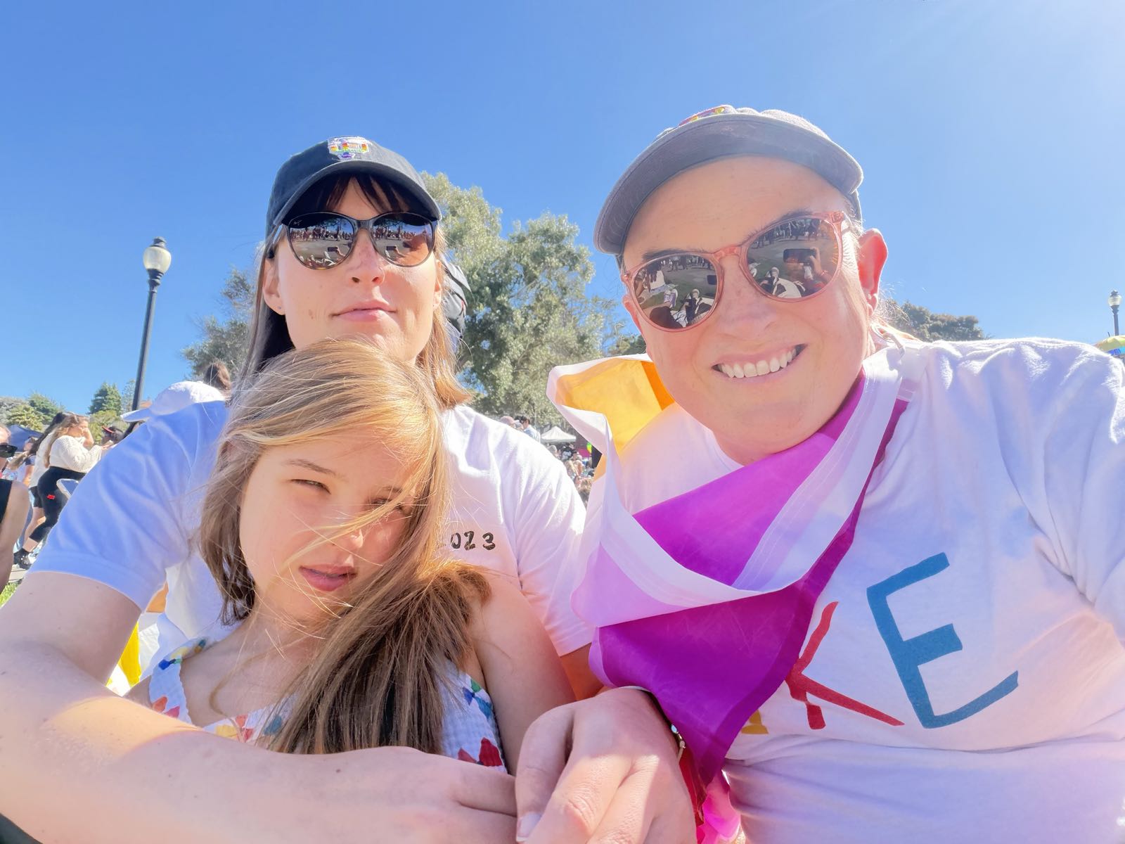 Two women and a girl pose for a photo on a bright summer day. Both women are wearing baseball hats and sunglasses.