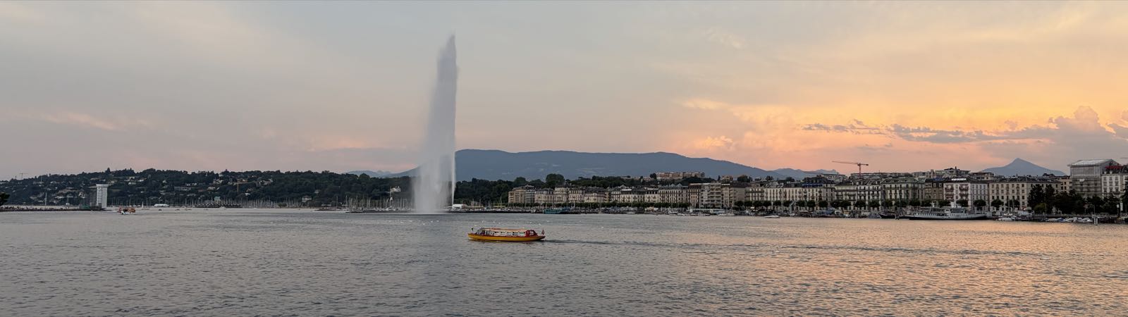 A yellow boat navigates the waters of a lake. Just beyond the boat, a large jet of water is being shot into the air from the center of the lake. There are many buildings aong the far shore, and mountains beyond them. It’s either sunrise or sunset, given the color of the sky.