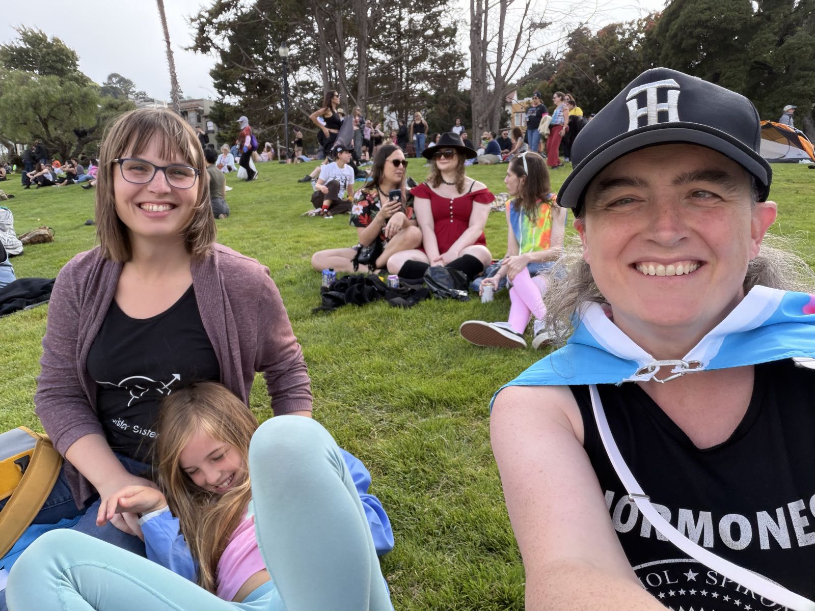 Two women and a girl sit in a park. The girl is leaning back onto one of the women, and is mid-laugh. Both women are smiling at the camera.