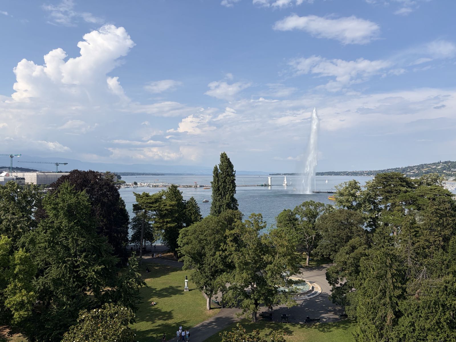 A park with trees is situated under a partly cloudy sky. There is a lake beyond the park, out of which a large jet of water is being sprayed.