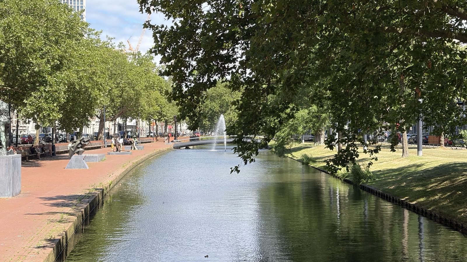 A canal is lined with trees and buildings. The right bank is grassy, and the left is made of brick. In the distance, there’s a fountain in the center of the canal.