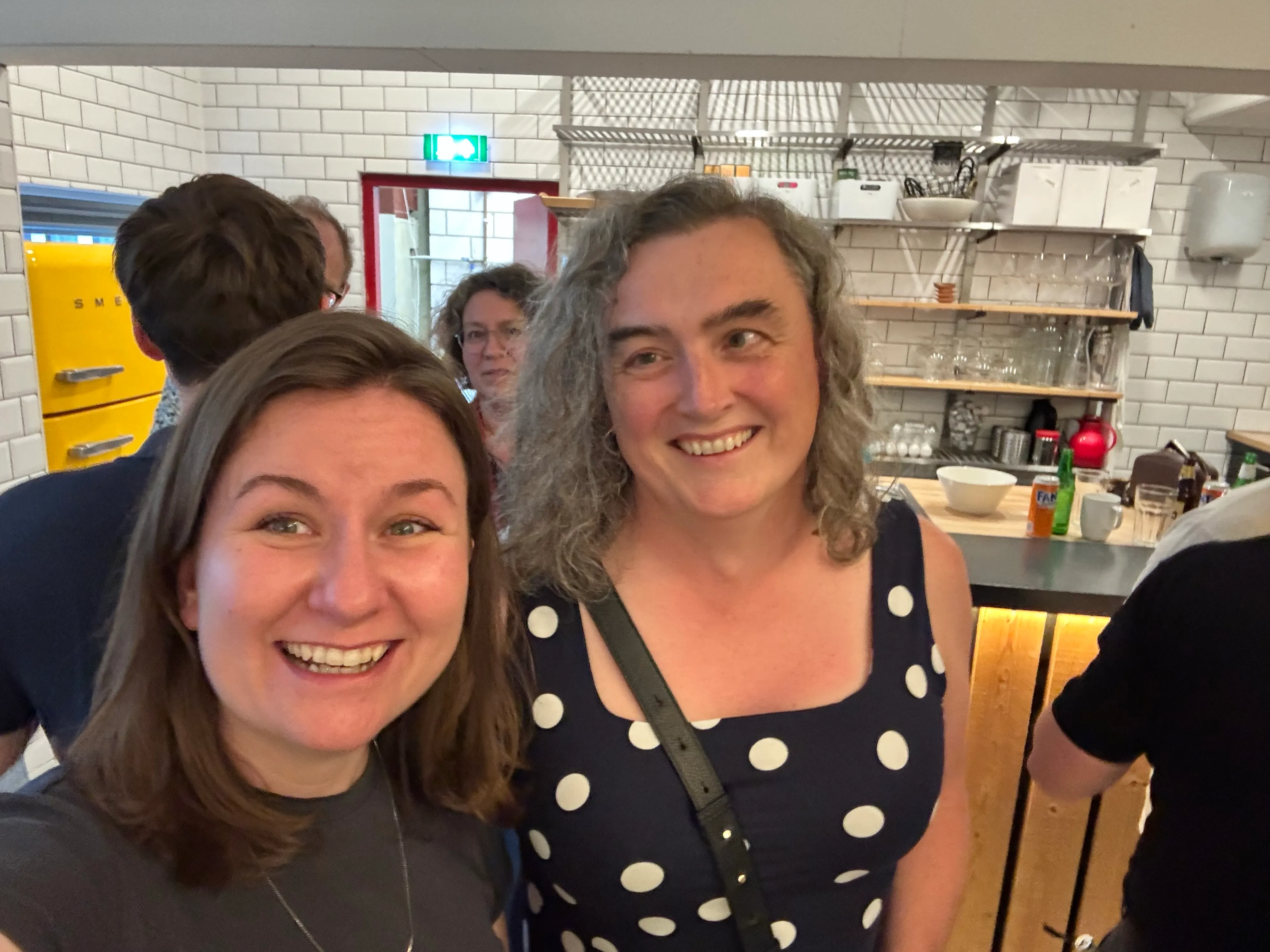 Two women pose for a selfie together. The one on the left has a gray t-shirt on, and the one on the right is in a navy dress with white polka dots.
