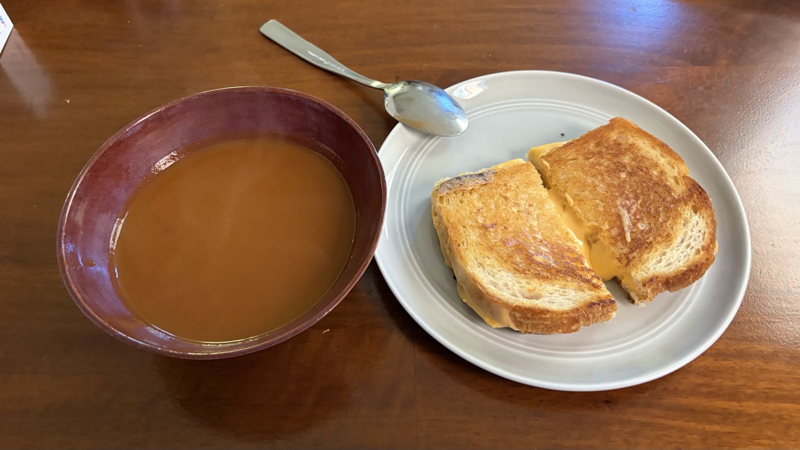 There’s a grilled cheese sandwich on a gray plate next to a red bowl filled with tomato soup.
