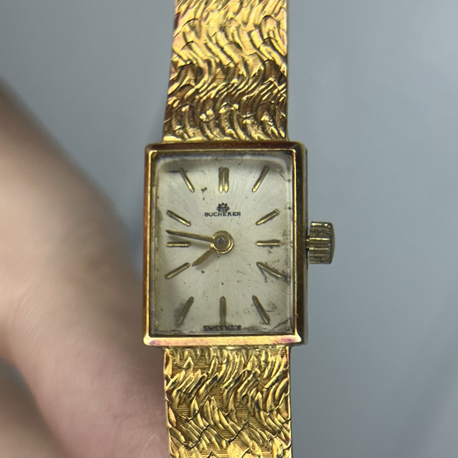 A small, rectangular, gold watch has a simple white dial with gold stick indicies and matching gold stick hands. Its gold bracelet has a wavy pattern.