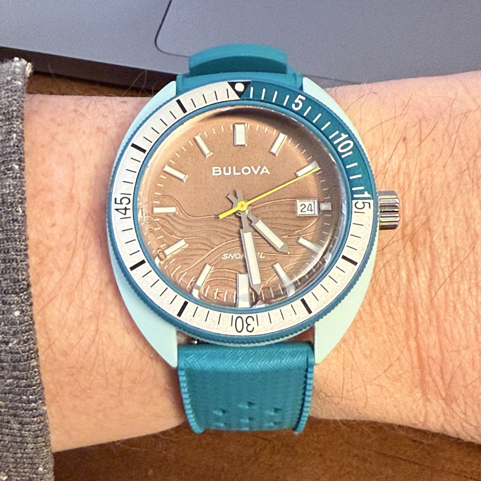 A mint-green watch with a blue-green rubber strap is on somebody’s wrist. It has a brown dial with a wave pattern on it. There’s a dive-style bezel with silver and green elements.