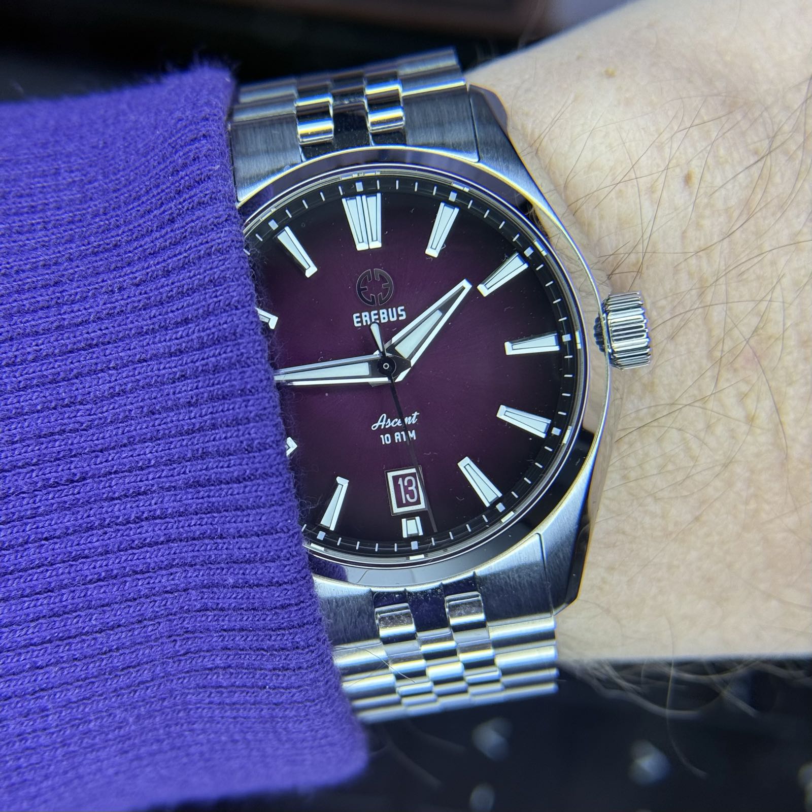 A shiny watch with a purple fumé dial is worn on the wrist of somebody in a purple sweatshirt.