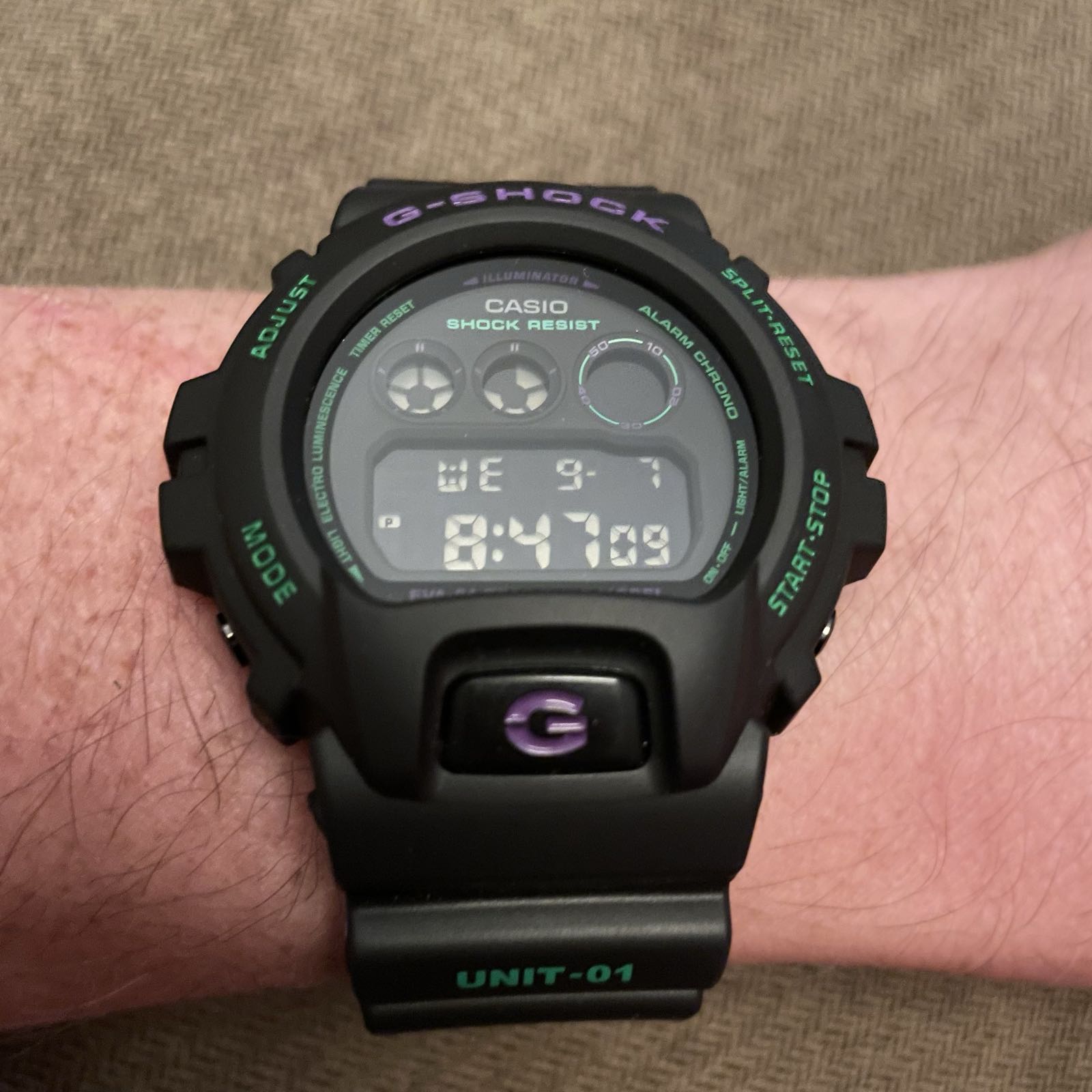 A black plastic digital watch with a black dial. The text on the bezel, dial, and strap is in green and purple. There are four positive displays: three circular displays in a row at the top, and a date and time readout at the bottom. Below the dial is a large black button with a purple G in the middle.