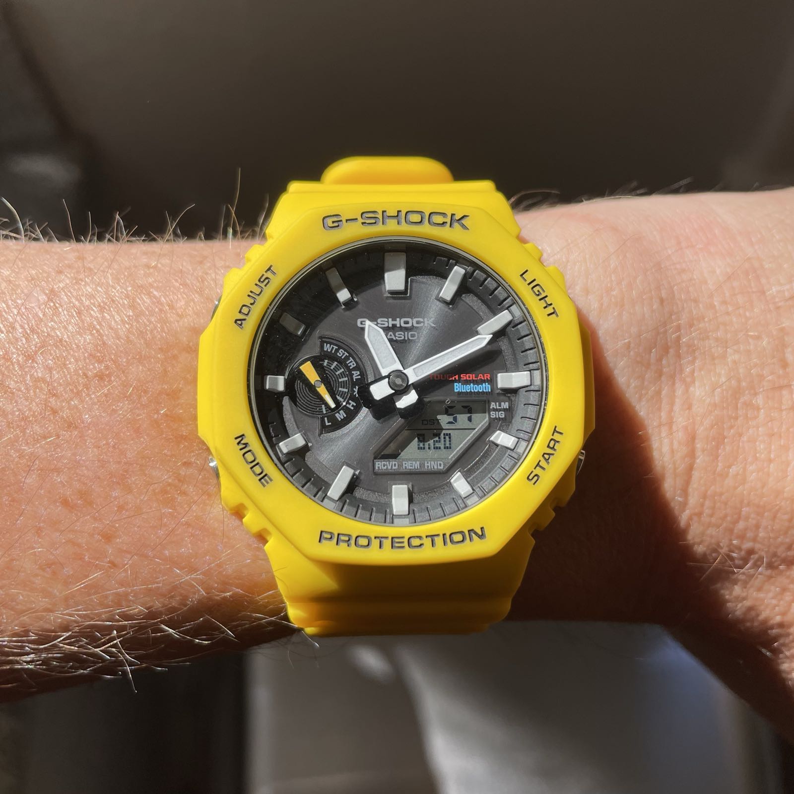 A yellow plastic analog/digital watch on a matching yellow plastic strap. The display is a dark gray, with a sunburst effect. The hour markers and hands are white.