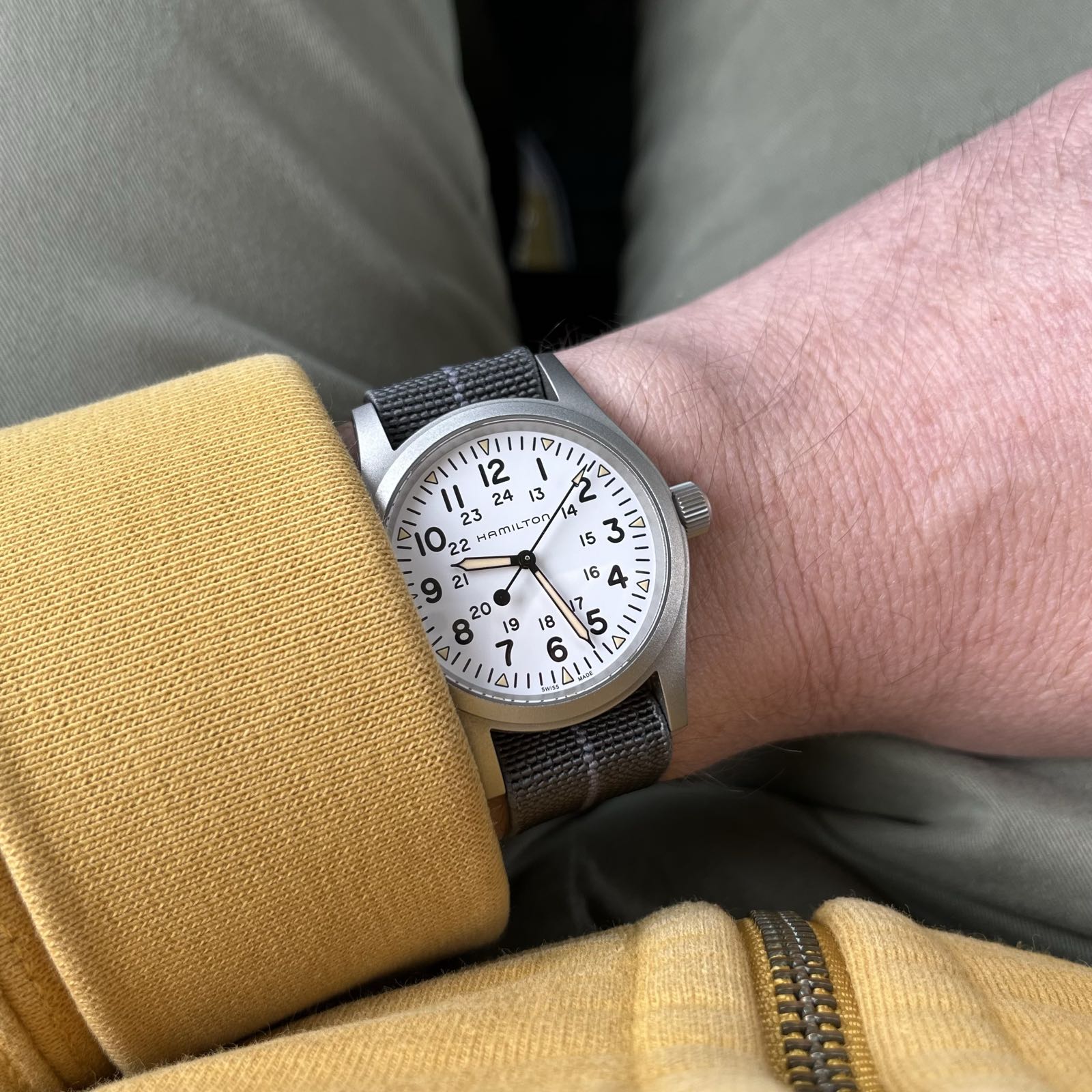 Someone in a yellow hoodie and khaki green pants is wearing a steel watch. The case finish is bead-blasted. The dial is stark white, with black arabics at each hour. The hands have a faux patina lume applied. The watch is on a gray nylon strap with a lighter gray centerline.