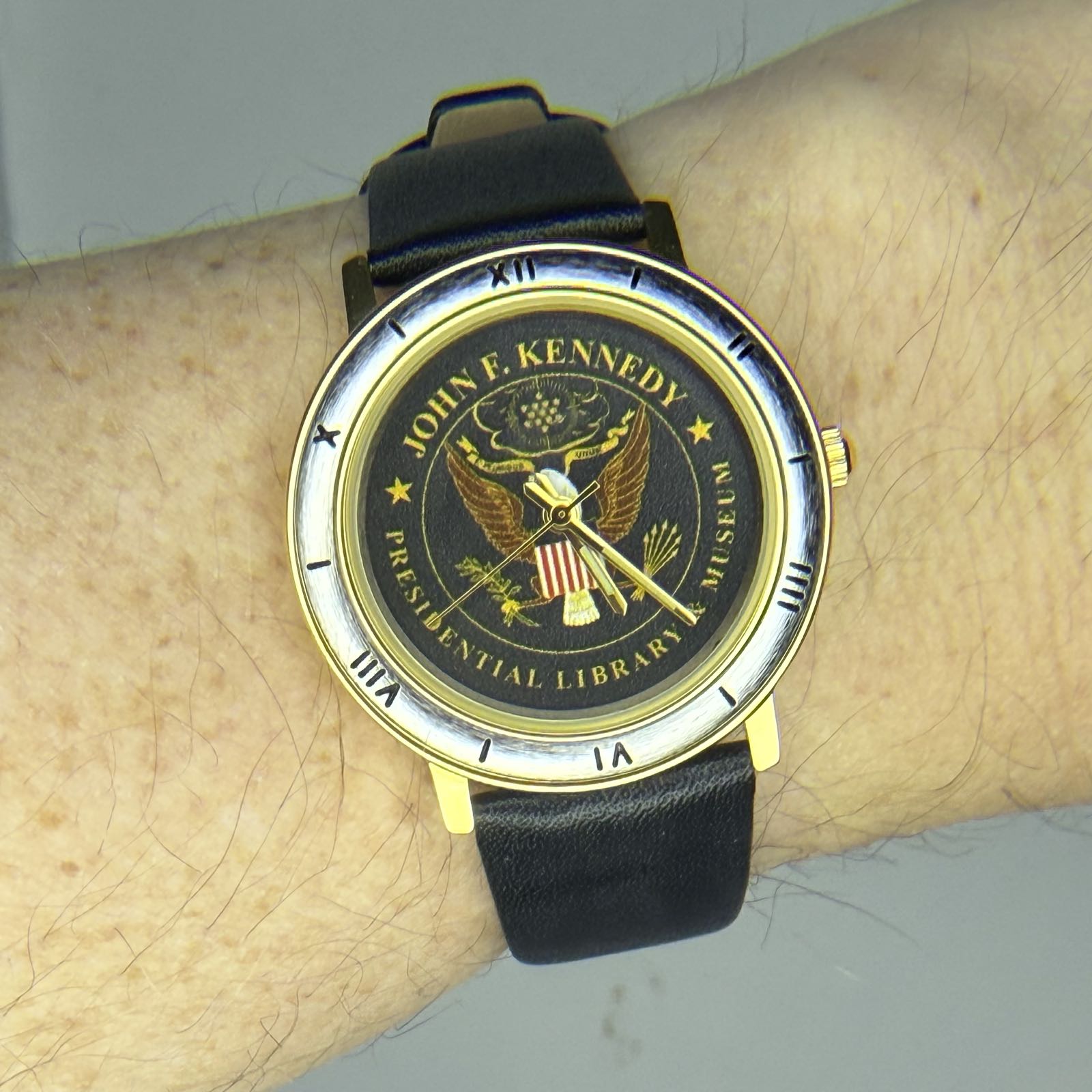 A gold-colored watch is being worn on a black leather strap. The watch has a blue dial, which features JFK’s presidential seal on it. The bezel is silver in color, and features a mixture of Roman numerals and stick indicies.