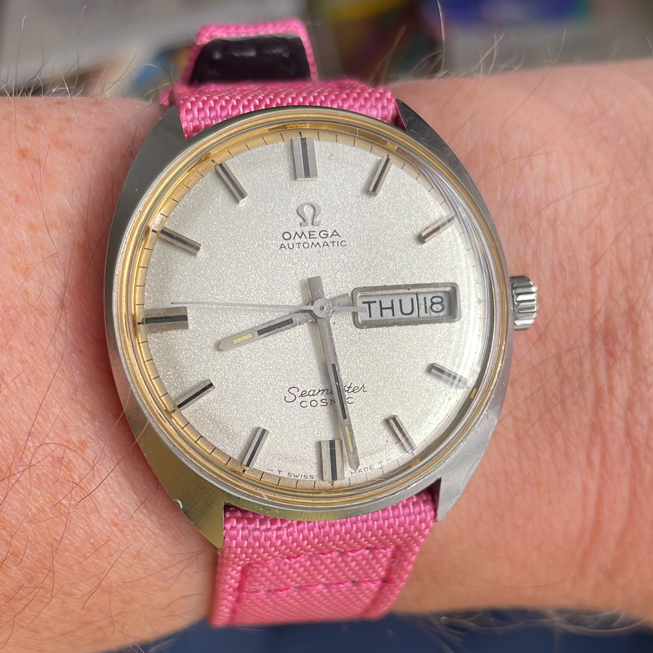A vintage Omega watch on a pink sailcloth strap. The watch has a cushion case, with simple stick indicies and hands on a sparkly silver dial. Theres a day-date complication at 3:00.