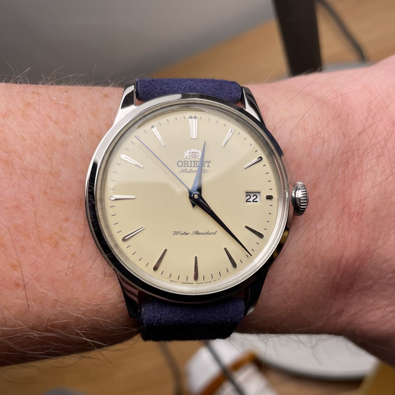 A simple, high-polish, steel dress watch with a champagne dial. The watch has blue hands and a date complication at 3:00. It’s on a navy blue leather strap.
