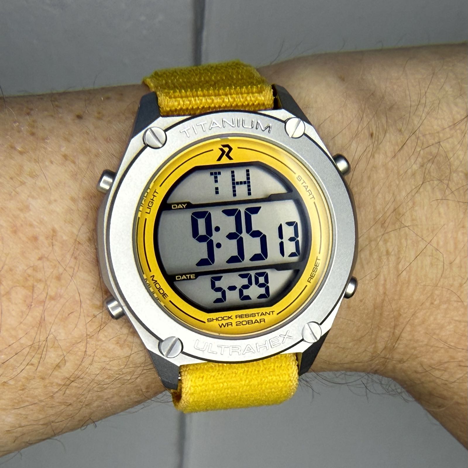 A metal watch with yellow details is on a yellow strap. The large display is digital, and very legible.