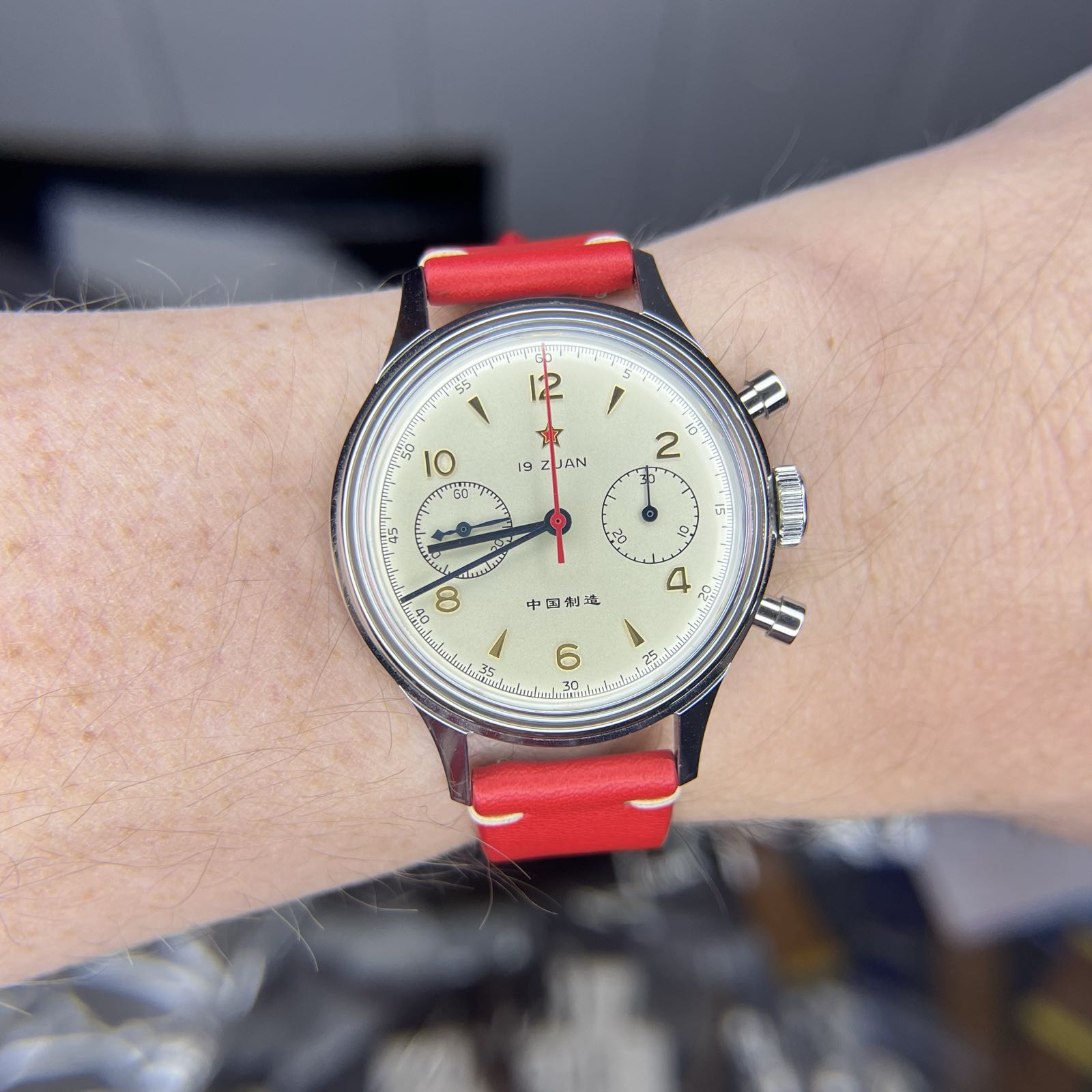A champagne-dial steel chronograph on a red leather strap. The watch has blue hour and minute hands, and blue hands on each of the two sub-dials at 3:00 and 9:00. The center chrono hand is red. Gold arabics are at the even hours, while arrow indicies are at the odds.