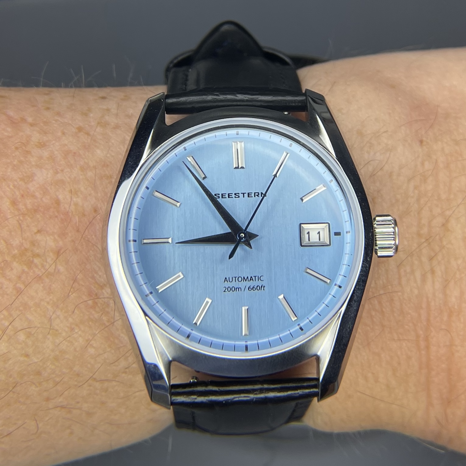 A steel dress watch on a navy blue leather strap. The case is high polish and has lyre lugs. The face is baby blue in color with vertical brushing throughout. Simple stick indicies mark the hours except for at 3:00, where there’s a date complication. The hour and minute hands are silver swords, while the needle seconds hand is blue.