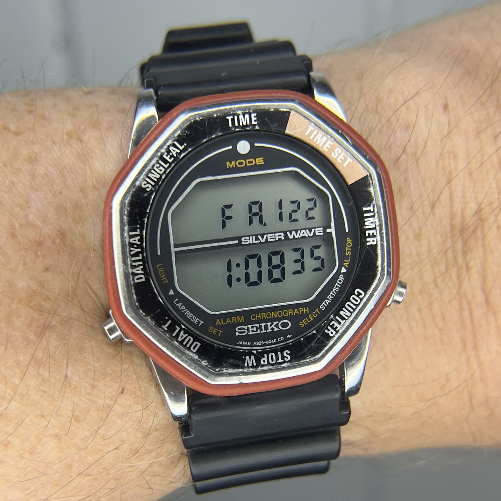 A steel digital watch on a black rubber strap. The positive digitial display is divided in two. The upper half displays the day and date, while the lower half displays the time. An octagonal rotating bezel surrounds the black dial. Each side of the bezel is labeled with a function. The upper right bezel segment is organge; the rest of the bezel segments are black. The bezel has a red rubber strip along the edge for easier gripping.