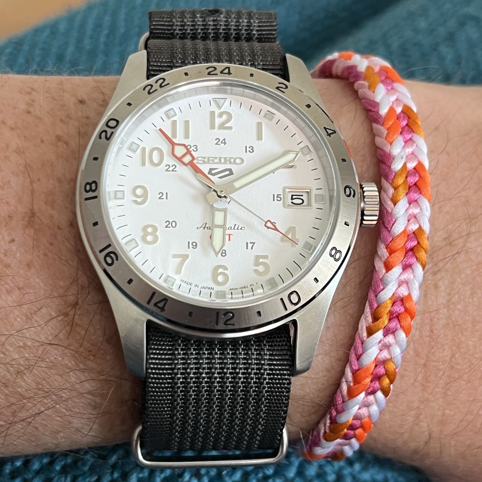 A steel watch with a white dial.
There’s a steel bezel with 24-hour markings around the watch’s crystal.
The dial is marked like a traditional field watch.
There’s an orange GMT hand which tracks a second time zone, and there’s a date complication at 3.
The watch is on a dark gray NATO strap.
