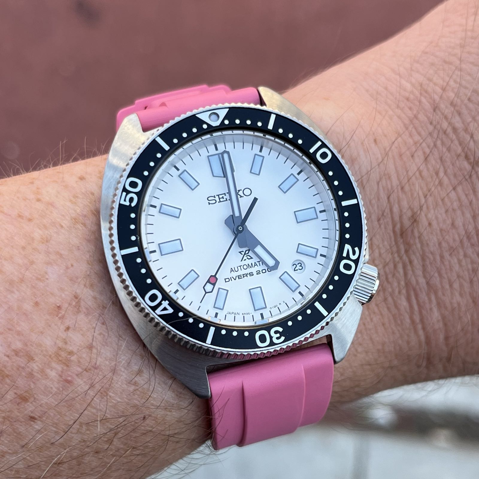 A steel, cushion-case dive watch on a pink rubber strap. The dial is white and the bezel insert is black. Stick indicies mark the hours, and there’s a circular date complication at 4:30. The tip of the seconds hand has one lumed section and one red section, resembling a stop light.