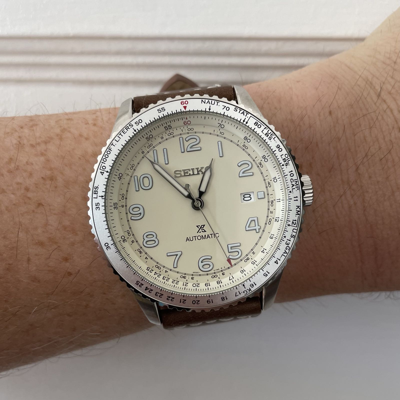 A large pilot’s watch on a brown leather strap. The watch has a champagne colored dial, simple stick hands, and large arabics at all of the hours except for 3:00, where it has a date complication. The aluminum bezel and the champagne chapter ring have slide rule markings, enabling the wearer to do a number of calculations.